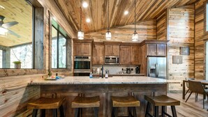 A rustic kitchen with wooden cabinets, a large island with stools, stainless steel appliances, and pendant lighting. - Modern and Luxury Cabin, 2 BR 2.5 Bath, Sleeps 9 max, 2 Pets Max with Pet Fee (Broken Bow)
