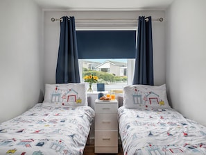 A bedroom with two beds, a window with curtains, a bedside table with a vase of flowers, and a view of a house outside.