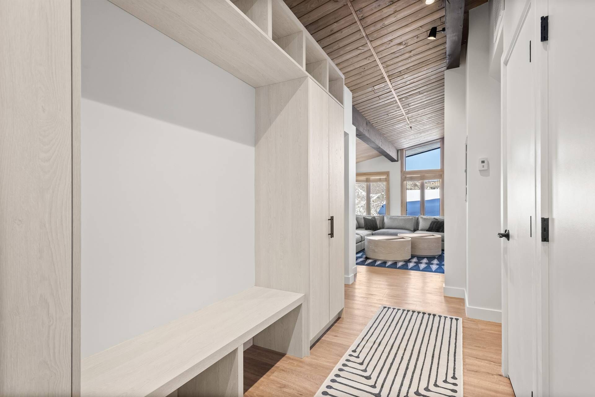 A hallway with a bench, a patterned rug, and a view into a living area with a sofa and cushions.