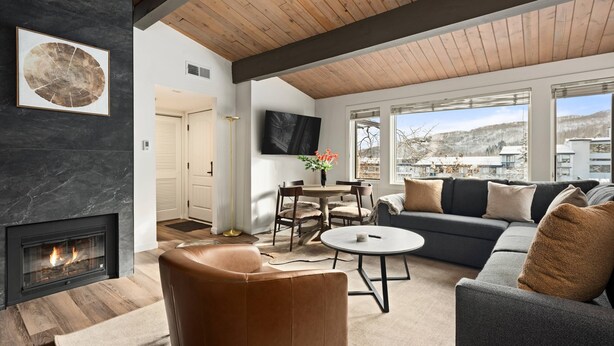 A modern living room with a fireplace, a large sofa, a dining area, and a TV mounted on the wall. - Aspenwood by iTrip Aspen Snowmass (Snowmass Village)