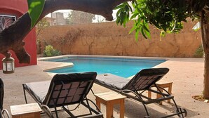 Une piscine avec deux chaises longues et une petite table, entourée d’un bâtiment rouge et d’un mur de briques.