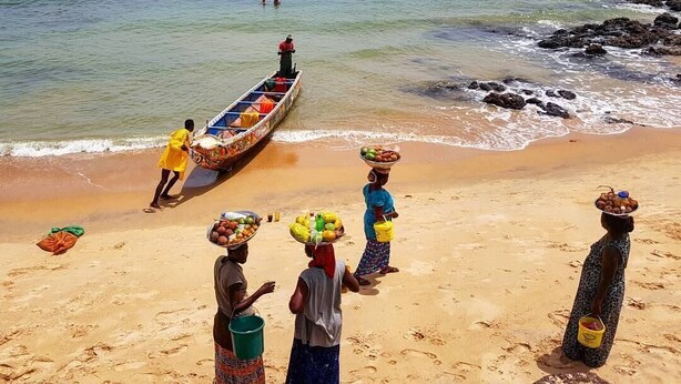 Une plage où l’on voit des personnes portant des marchandises sur leur tête, et un bateau sur l’eau.