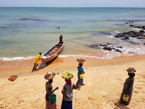 Une plage où l’on voit des personnes portant des marchandises sur leur tête, et un bateau sur l’eau.