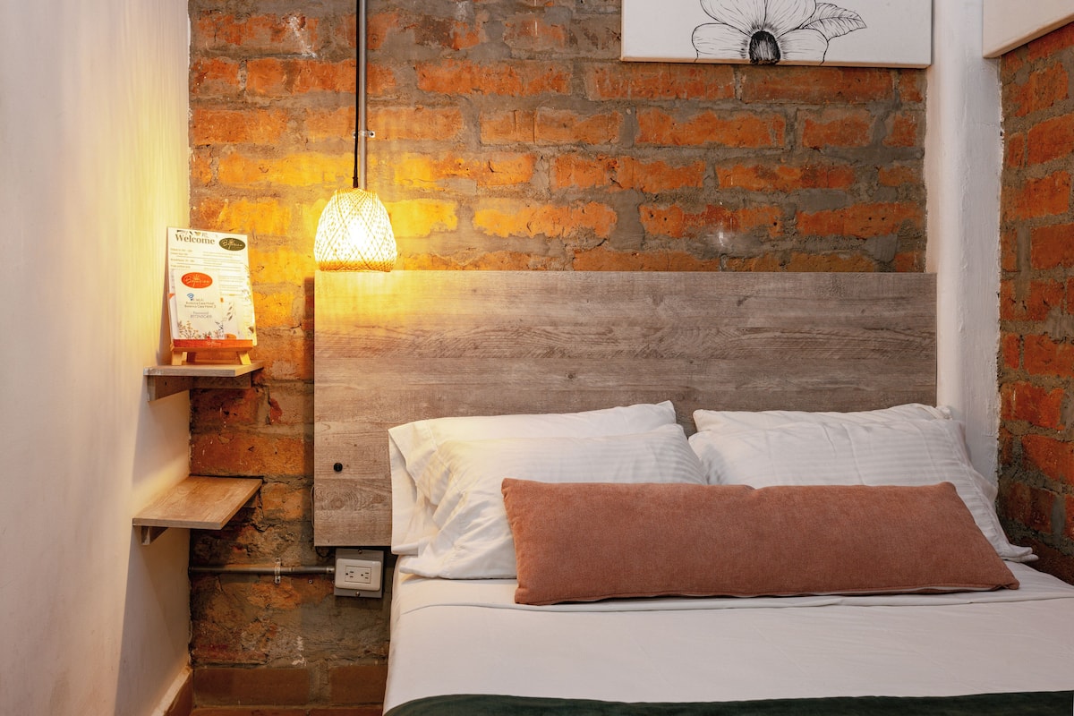 Una cama con sábanas blancas y un almohadón marrón, un cabecero de madera, una pared de ladrillo, una lámpara colgante y un letrero de bienvenida.