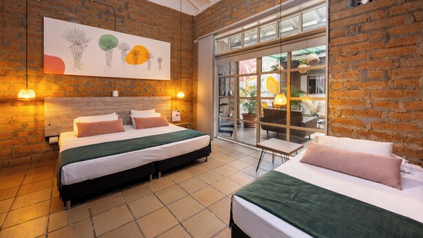 A hotel room with a brick wall, a large window, and a bed with a green bedspread. - Botánica Casa Hotel (Medellín)