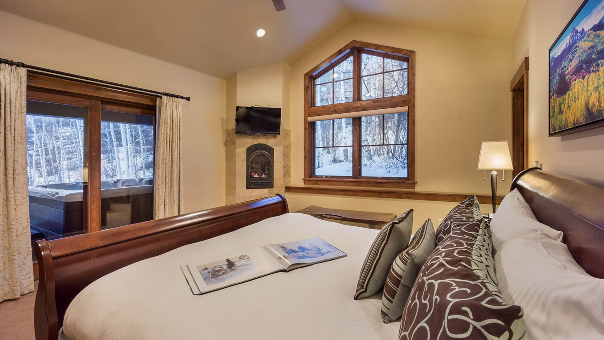 A hotel room with a large bed, a fireplace, a large window with a view of trees, and a painting on the wall.