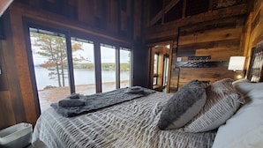 Een slaapkamer in een houten blokhut met een bed, uitzicht op een meer en een houten plafond.