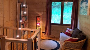 En mysig stuga med trägolv, en trappa, en bokhylla, ett fönster med gardiner och en soffa med en kudde.