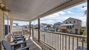 A covered porch with wooden chairs and tables, a white railing, and a view of residential houses.