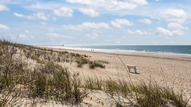 A beach with sand dunes, a bench, and the ocean.