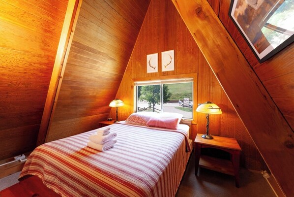 A cozy, wooden-paneled room with a bed, two bedside lamps, and a window with a view of the outside. - CJ A-Frame 2 - In Town - Ski In/Ski Out - On The River - Free WiFi (Red River)