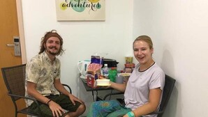 Twee personen zitten aan een tafel met diverse gerechten en een koffieapparaat, in een ruimte met een deur en een muur met een bord.