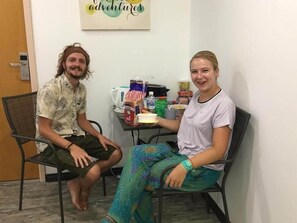 Two people seated at a table with various food items and a coffee machine, in a room with a door and a wall with a sign. - Signel Hostel (Kota Kinabalu)