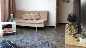 Een woonkamer met een beige bank, een klein wit tafeltje met een stapel kleurrijke blokken, een pluizig grijs tapijt en een televisie op een houten dressoir.