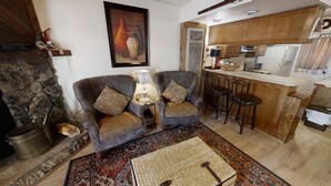 Un salon comprenant deux fauteuils, une table basse, une cheminée et un coin cuisine avec des meubles en bois et des tabourets de bar.
