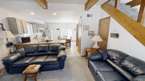 A living room with a sofa, a coffee table, and a lamp. There is a kitchen area with cabinets and appliances. A wooden staircase is visible in the background. - LRM Cabin - In Town - Walk to Main Street - WiFi - Satellite - Wood Burning Fire (Red River)