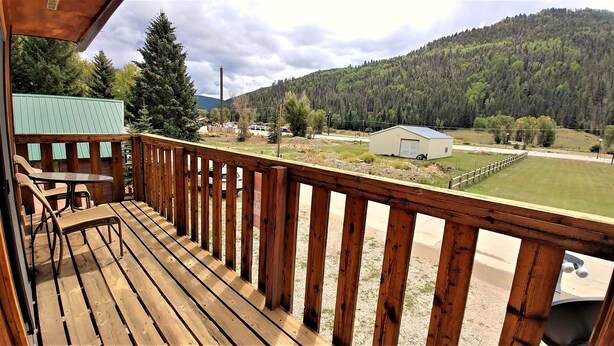 A wooden deck with a table and chairs, overlooking a grassy area with a small building and a forested hill in the background. - LRM Cabin - In Town - Walk to Main Street - WiFi - Satellite - Wood Burning Fire (Red River)