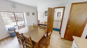 Un área de comedor con una mesa y sillas de madera, una ventana con vistas a árboles, y una puerta que da a una cocina.