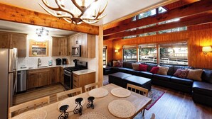 Una cocina moderna con armarios de madera, un refrigerador de acero inoxidable y una lámpara de araña grande. Hay un comedor con una mesa y sillas de madera, y una zona de estar acogedora con un sofá y cojines.