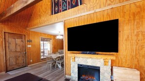 Interior acogedor de una cabaña con una chimenea de piedra, un televisor montado encima y una zona de comedor con una mesa y sillas.