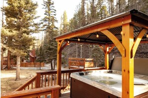 Een houten jacuzzi onder een overdekte veranda met een houten terras en banken, omringd door bomen.
