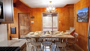 Comedor con paneles de madera, una mesa dispuesta para seis comensales, una araña y una ventana con persianas.