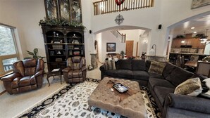 A spacious living room with a large sectional sofa, leather armchairs, and a coffee table. There is a fireplace with a mantle, a bookshelf filled with books, and a staircase leading to an upper level.
