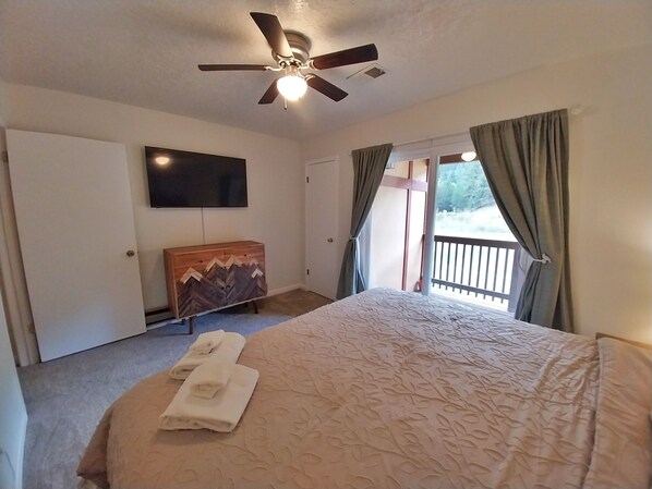 A hotel room with a bed, a ceiling fan, a TV mounted on the wall, a wooden dresser, and a balcony with curtains.