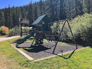 Children can play safely within the enclosed playground area, featuring a slide and swing set, surrounded by lush greenery and a well-maintained grassy space.
