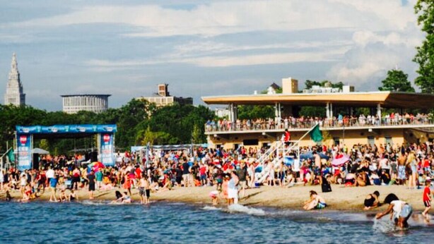 En välbesökt strand med folk som badar och promenerar längs strandkanten, en stor blå portal och byggnader i bakgrunden.