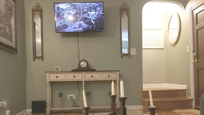 Ett rum med en tv som hänger på väggen, en klocka, en ljusstake och en trappa som leder upp till ett övre våningsplan.