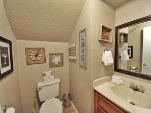 Ein Badezimmer mit Toilette, Waschbecken und Spiegel. An der Wand hängen Bilder in Rahmen und ein Mülleimer steht bereit.