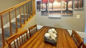 A dining area with a wooden table, chairs, a staircase, and three autumn-themed paintings on the wall.