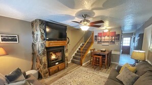 A living room with a stone fireplace, a television mounted above it, a ceiling fan, a staircase leading to another level, and a dining area with a table and chairs.