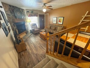 Eine Treppe führt zu einer zweiten Etage mit Holzwertung, einem Wohnzimmer mit Sofa und Kamin, sowie einem Deckenventilator.