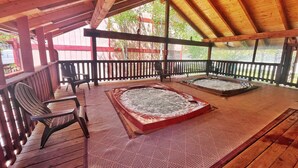 A wooden deck with two hot tubs, a chair, and a view of greenery.