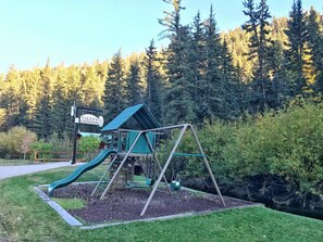 A playground with a slide and swing set, surrounded by a forested area.