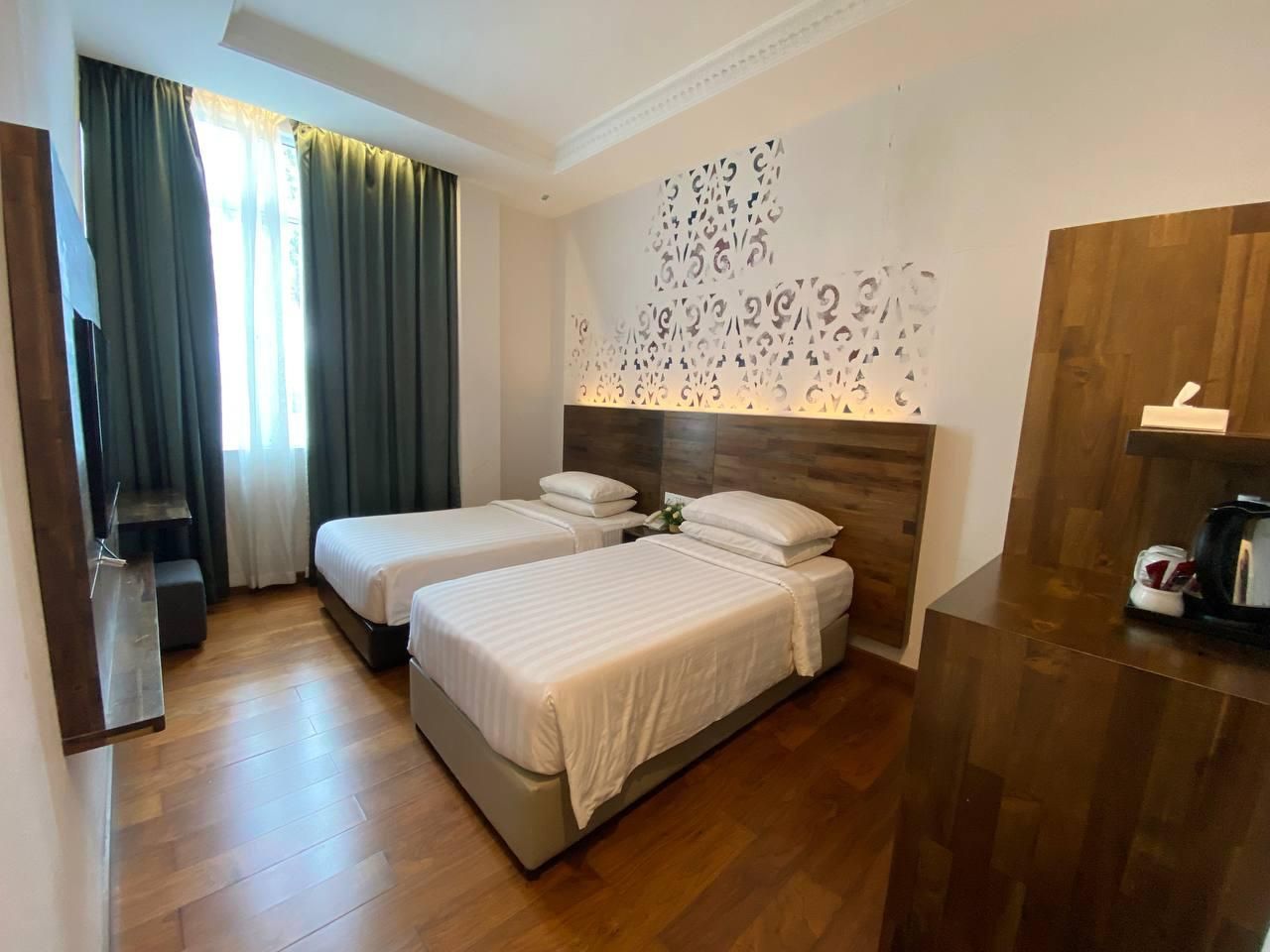 A hotel room with two beds, a wooden floor, a wall-mounted decorative panel, and a wooden desk with a chair.