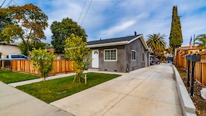 A residential street with a single-story house, a well-maintained lawn, and a clear sky.