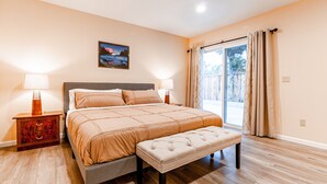 A bedroom with a large bed, two bedside tables with lamps, a bench at the foot of the bed, a window with curtains, and a framed picture on the wall.