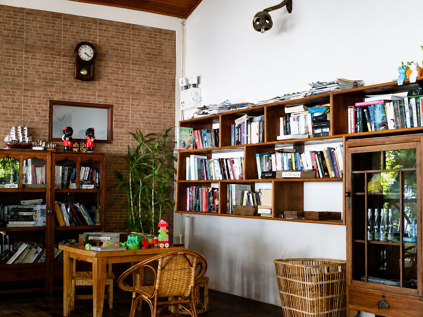 A cozy reading nook with a brick wall, a wooden bookshelf filled with books, a small table with a chair, and a decorative clock.