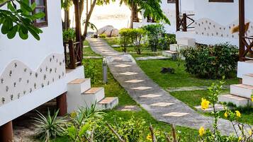 A pathway leading to a building with a white exterior and a red roof, surrounded by greenery and a clear view of the sea.