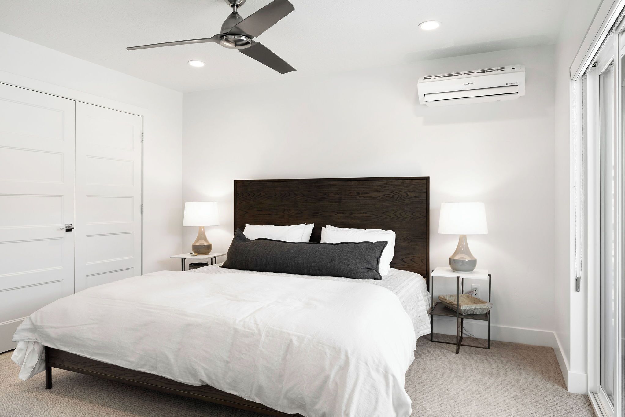 Une chambre à coucher avec un lit, deux tables de chevet avec des lampes, un ventilateur de plafond, un appareil de climatisation et une porte blanche.