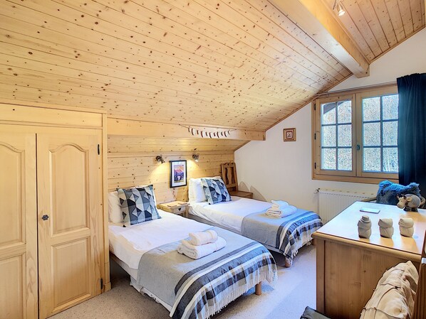 A cozy room with two beds, wooden ceiling, a window with a view, and a small table with decorative items.