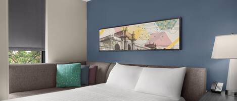 A hotel room with a bed, a nightstand with a lamp, a painting on the wall, and a window with a view of trees.