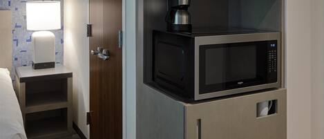 Habitación de hotel compacta con kitchenette, que incluye microondas y cafetera, una lámpara de noche y un papel tapiz estampado.