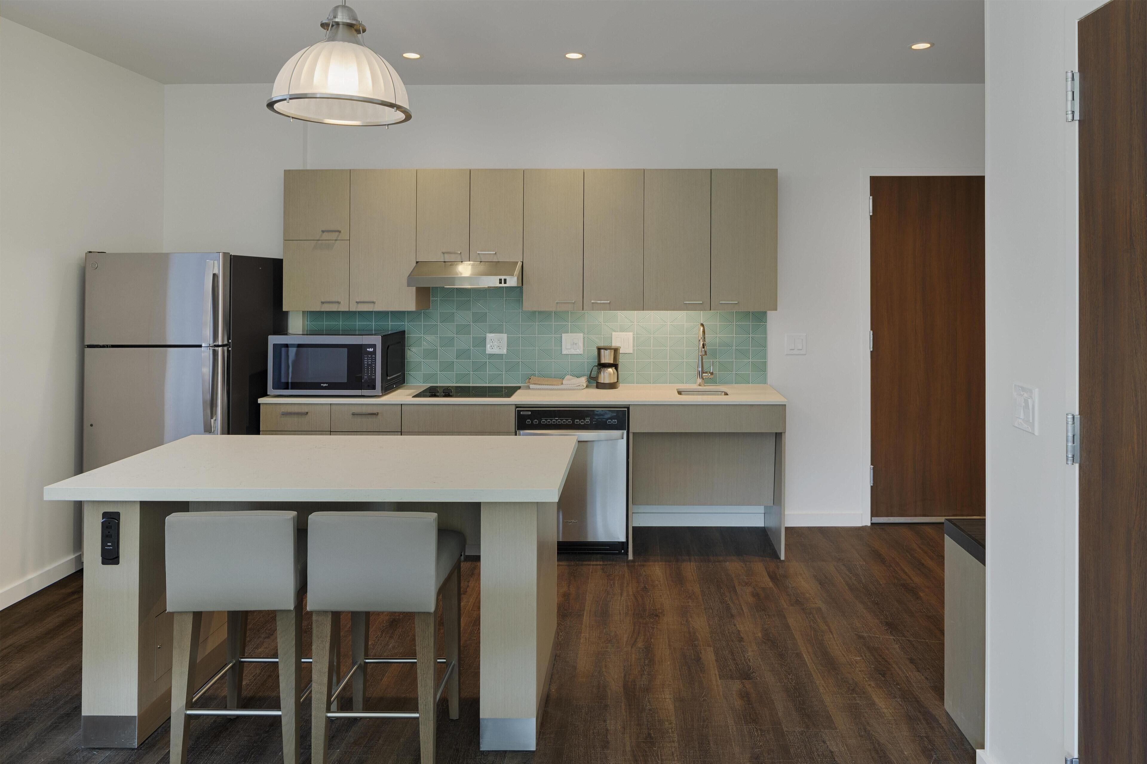 A modern kitchen with a stainless steel refrigerator, microwave, and dishwasher, a wooden dining table with two chairs, and a wooden door.
