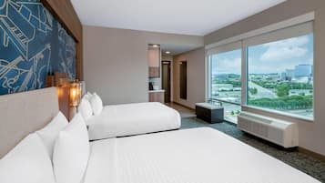 A hotel room with two beds, a large window with a city view, a wall-mounted lamp, and a radiator.