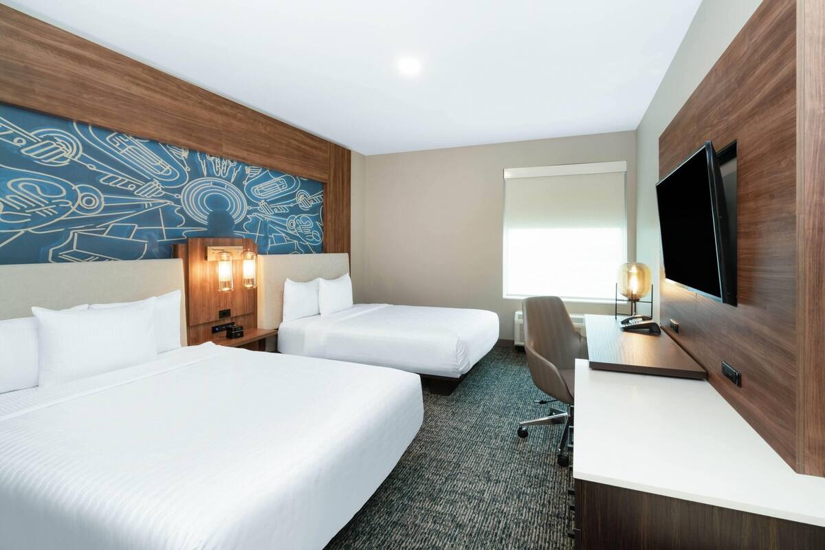 A hotel room with two beds, a desk, a TV, and a wall with a blue abstract design.