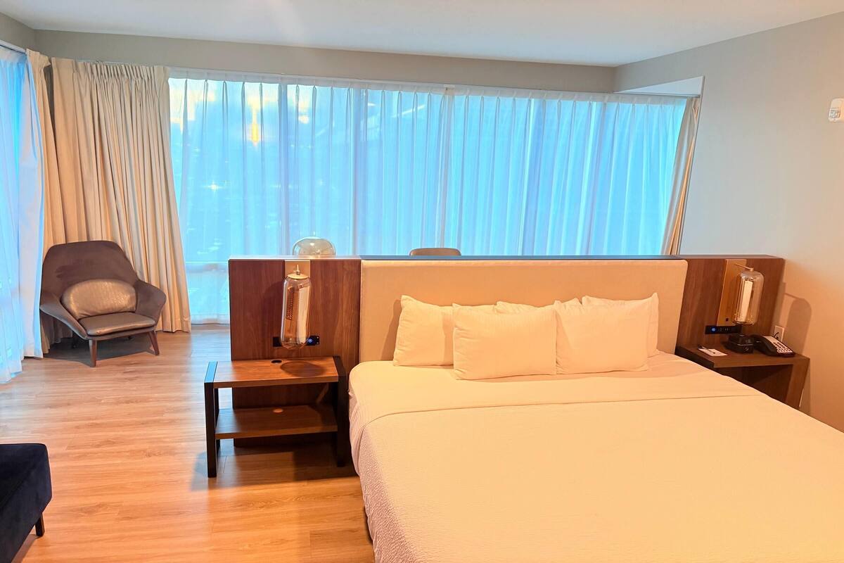 A hotel room with a large bed, a wooden nightstand, a lamp, a chair, and a view of the city through sheer curtains.